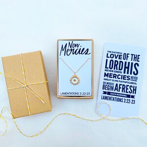 May include: A gold necklace with a sun pendant and a card that reads "The faithful love of the Lord, His mercies never cease. Great is His faithfulness; His mercies begin afresh each morning. Lamentations 3:22-23". The necklace is in a small white box with the words "New Mercies" and "Lamentations 3:22-23" printed on it.