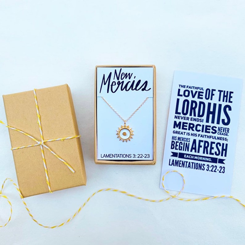 May include: A gold necklace with a sun pendant and a card that reads "The faithful love of the Lord, His mercies never cease. Great is His faithfulness; His mercies begin afresh each morning. Lamentations 3:22-23". The necklace is in a small white box with the words "New Mercies" and "Lamentations 3:22-23" printed on it.