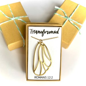 May include: A gold necklace with a butterfly pendant and the words "Transformed" and "Romans 12:2" on a white card in a brown box.