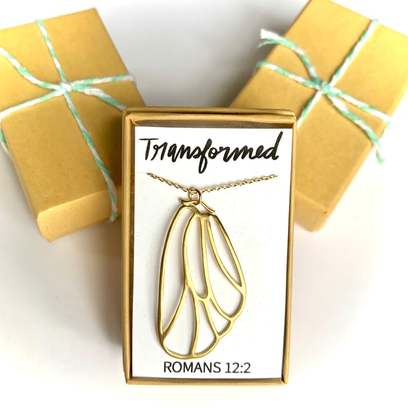 May include: A gold necklace with a butterfly pendant and the words "Transformed" and "Romans 12:2" on a white card in a brown box.