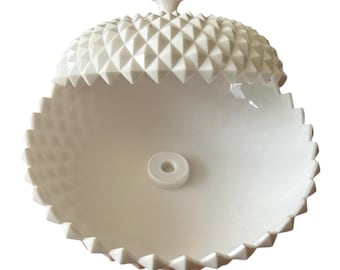 May include: A white, decorative lidded bowl with a textured, geometric pattern. The bowl's lid and base feature a repeating diamond design, and the rim has a jagged edge. A small, circular piece is in the center of the bowl.