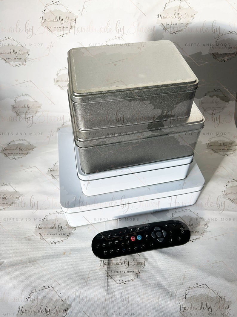 May include: A stack of four silver and white metal tins with a black remote control on a white background with a repeating pattern of black and gray geometric shapes and the text "Handmade by Stacey Gifts and More".