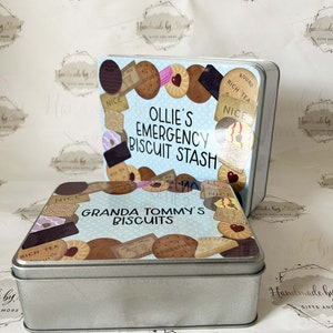 May include: Two silver metal biscuit tins with blue and white lids. The top tin has the text "Ollie's Emergency Biscuit Stash" and the bottom tin has the text "Granda Tommy's Biscuits". Both tins are decorated with images of various biscuits.
