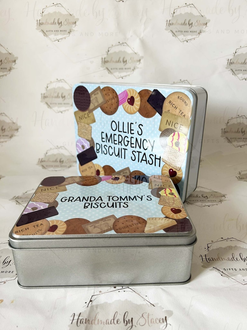 May include: Two silver metal biscuit tins with blue and white lids. The top tin has the text "Ollie's Emergency Biscuit Stash" and the bottom tin has the text "Granda Tommy's Biscuits". Both tins are decorated with images of various biscuits.