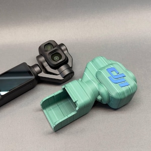 Op de afbeelding: Een zwarte handheld gimbal-camera met een dubbel lenssysteem naast een lichtgroene 3D-geprinte beschermhoes. De hoes heeft een getextureerd, gelaagd oppervlak en een opvallend blauw DJI-logo op het bovenste gedeelte.