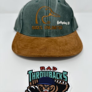 Peut inclure: Casquette de baseball Ducks Unlimited verte et marron avec une visière marron. La casquette présente un logo de canard brodé en or et le texte "Ducks Unlimited" et "Gettysburg, SD". Un autocollant avec un ours portant une casquette bleue et le texte "RAD THROWBACKS" est en dessous.