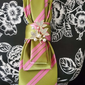 May include: A necktie featuring a diagonal striped pattern in olive green and bright pink. The tie is folded and secured with a gold-toned brooch shaped like a leafy branch, embellished with clear crystals and small white faux pearls.