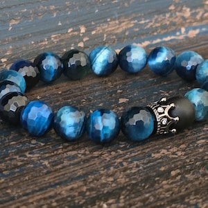 May include: A beaded bracelet featuring faceted blue tiger eye stones and a black bead with a silver crown accent. The bracelet is strung on an elastic band and rests on a distressed wooden surface.