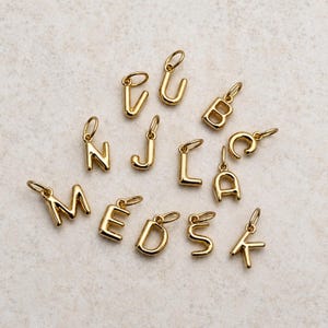May include: A collection of gold-toned metal letter charms, each featuring a small circular jump ring at the top for attachment. The set includes individual alphabet characters with a smooth, polished finish, arranged in a scattered layout.