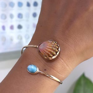 May include: A gold-toned open cuff bracelet featuring two distinct focal points: one end displays a textured, fan-shaped seashell with warm orange and yellow hues, while the other end holds a smooth, oval-shaped light blue stone with white marbling.