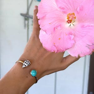 May include: A metal cuff bracelet featuring two distinct ends: one end is a small, white, cone-shaped shell with black spots, and the other end is a smooth, round, bright turquoise stone. The bracelet is worn on a wrist next to a large, vibrant pink hibiscus flower.