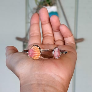 May include: A silver-toned open cuff bracelet featuring two distinct seashell accents. One shell is a larger, fan-shaped scallop with orange and pink gradients, while the other is a smaller, oval-shaped shell with pink and white vertical ridges.