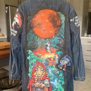 May include: A dark blue denim jacket with colorful artwork depicting a full moon, trees, and a Little Red Riding Hood scene. The jacket has the text "Ultimate Woman" and "Hey the rid you lo" on it.