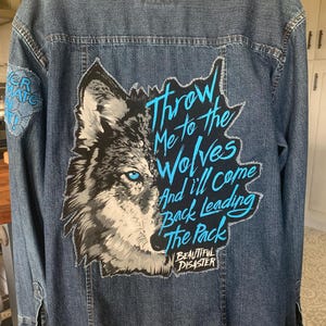 May include: A blue denim jacket with a wolf graphic and the text "Throw Me to the Wolves And I'll Come Back Leading The Pack". The wolf has blue eyes and is partially black and white.