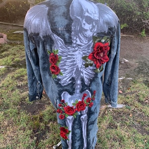 May include: A denim jacket with a skeleton design on the back, featuring a skull and rib cage, accented with red rose embroidery. The jacket has a distressed look with frayed edges, and is displayed on a mannequin.