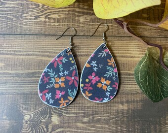 May include: A pair of teardrop-shaped earrings with a floral print. The earrings are black with pink, orange, and yellow flowers. The earrings have silver hooks.