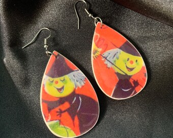 May include: A pair of dangle earrings featuring a vintage illustration of a smiling witch with a broom and cauldron. The earrings are made of wood and have a teardrop shape. The witch is depicted in a red, yellow, and green color scheme.