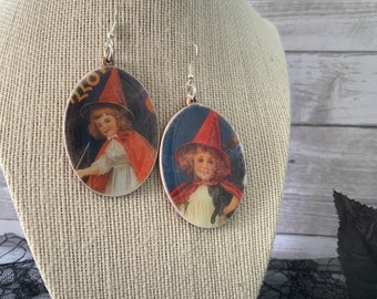 May include: A pair of silver dangle earrings with round, colorful images of a girl dressed as a witch. The images are printed on a white background and feature a black cat and a wand.