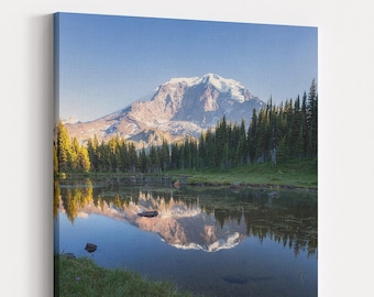 May include: A scenic landscape photograph featuring a mountain reflected in a still lake. The mountain is snow-capped and surrounded by lush green trees. The lake is calm and clear, with a blue sky reflecting in the water.