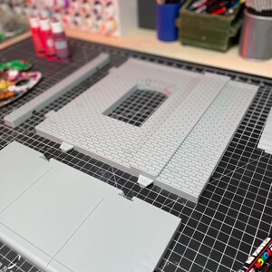 May include: A gray, multi-piece model of a building with a brick facade. The model is on a black cutting mat with a ruler and markers.