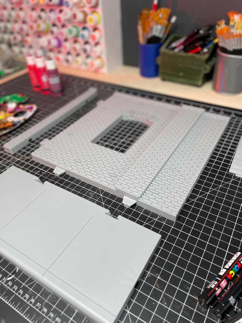 May include: A gray, multi-piece model of a building with a brick facade. The model is on a black cutting mat with a ruler and markers.