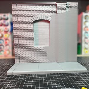 May include: A gray brick wall with a window and a sidewalk. The wall is made of a textured material and has a realistic look. The window is arched and has a gray frame. The sidewalk is made of concrete and has a smooth surface.