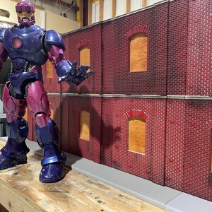 May include: A purple and silver action figure of a superhero standing in front of a red brick wall with boarded up windows. The figure has a helmet with a gold visor and a detailed suit.