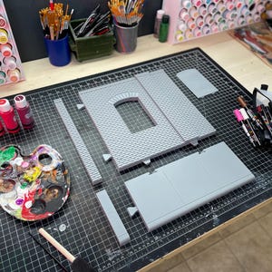 May include: A set of gray plastic model building pieces for a miniature structure, laid out on a black cutting mat. The pieces include a brick wall with an arched window, a flat roof section, and several straight wall sections. There are also paintbrushes, paint bottles, and a paint palette in the image.