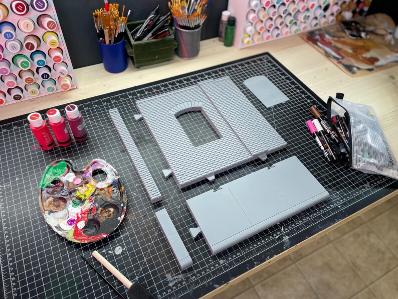 May include: A set of gray plastic model building pieces for a miniature structure, laid out on a black cutting mat. The pieces include a brick wall with an arched window, a flat roof section, and several straight wall sections. There are also paintbrushes, paint bottles, and a paint palette in the image.