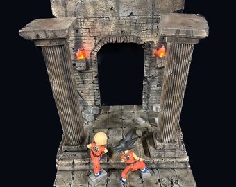 May include: A 1:12 scale diorama of a ruined stone archway with two figures standing in front of it. The archway is made of gray and brown stone and has a textured surface. The figures are wearing orange and black clothing. The diorama is lit by two small red lights.