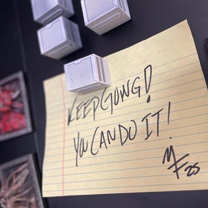 May include: A collection of white miniature air conditioner magnets are arranged on a black surface. A yellow notepad with lined paper is attached, featuring the handwritten message "KEEP GOING! YOU CAN DO IT!" and a signature.