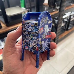 May include: A miniature blue mailbox with numerous stickers covering its surface. The mailbox has a curved top and four slender legs. The stickers vary in size and design, adding a personalized touch to the small, decorative item.