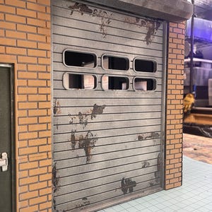 May include: A miniature garage model featuring a weathered, corrugated metal door with rectangular windows. The door is set between brick walls. The metal shows signs of rust, contributing to the aged appearance of the structure.