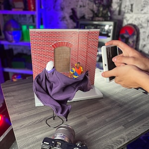 May include: A purple cape is draped over a stand in front of a brick building backdrop. A figure wearing a yellow shirt and a red hat is standing in the doorway of the building. A camera is positioned in front of the cape and a smartphone is being held in the right hand of a person taking a photo.