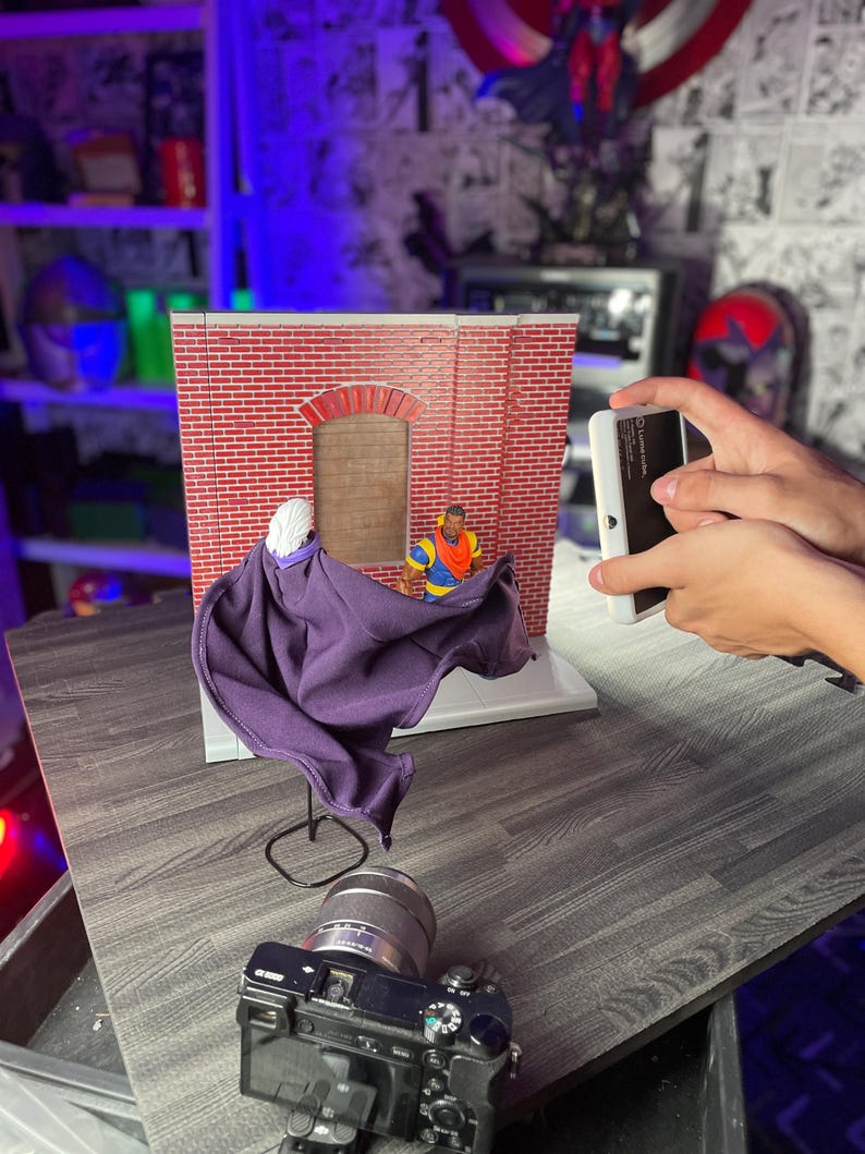 May include: A purple cape is draped over a stand in front of a brick building backdrop. A figure wearing a yellow shirt and a red hat is standing in the doorway of the building. A camera is positioned in front of the cape and a smartphone is being held in the right hand of a person taking a photo.