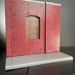 May include: A red brick wall with a gray concrete sidewalk in front. The wall has a single arched doorway with a wooden door. The wall is made of small bricks and has a rough texture.