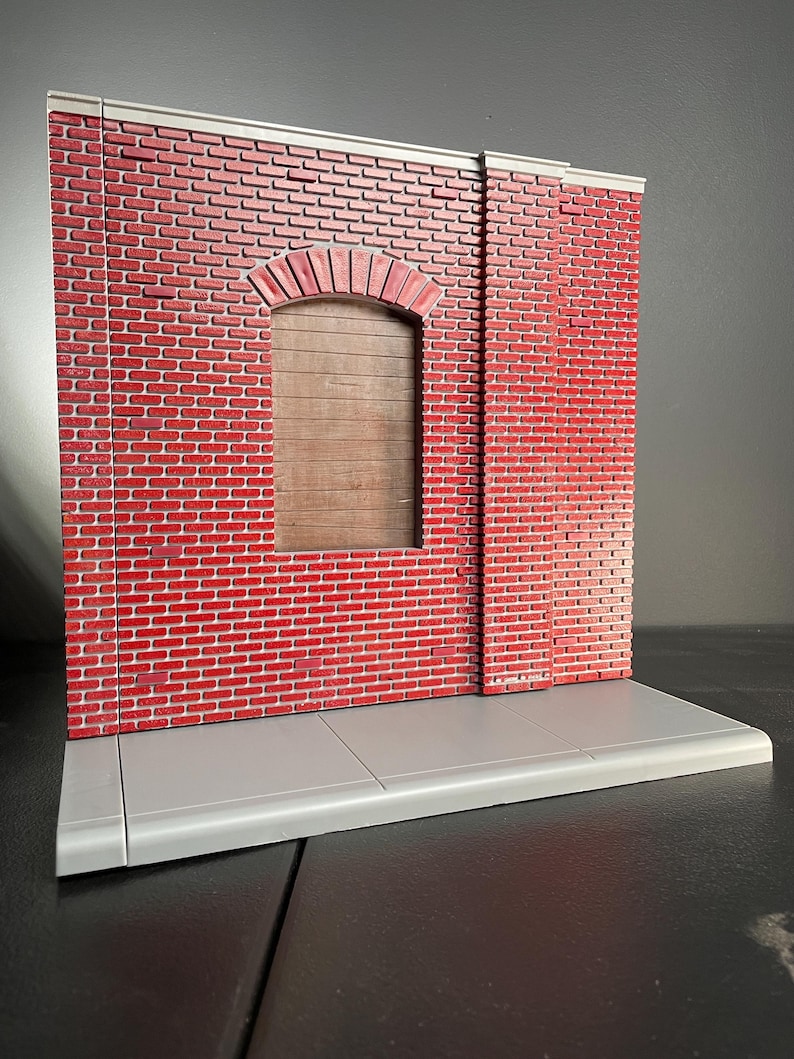 May include: A red brick wall with a gray concrete sidewalk in front. The wall has a single arched doorway with a wooden door. The wall is made of small bricks and has a rough texture.