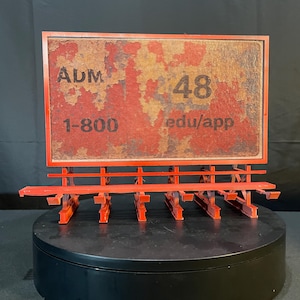 May include: A miniature red billboard with a weathered, textured sign. The sign displays the text "ADM 48" and a phone number and website address. The billboard is mounted on a black circular base.