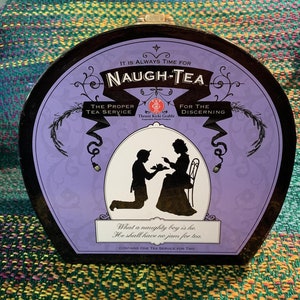 May include: A round, black tea tin with a purple lid. The lid features a whimsical illustration of a boy and a girl in silhouette, with the text "It is always time for Naugh-Tea. The Proper Tea Service. Thrusti Kicki Grabbi. For the Discerning. What a naughty boy is he. He shall have no jam for tea. Contains one tea service for two."