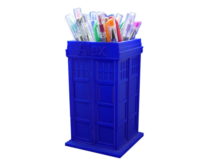 May include: Blue plastic pencil holder shaped like a police box with the name "Alex" on the front. The holder is filled with colorful pens and pencils.