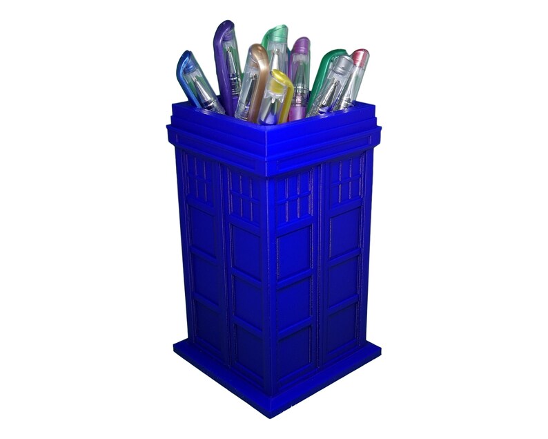 May include: A blue TARDIS-shaped pencil holder with colorful pens sticking out of the top.