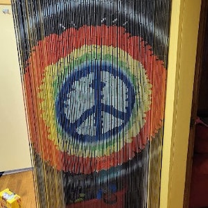 May include: A beaded curtain with a rainbow peace sign design. The curtain is made of black, vertical strands with a large, colorful peace symbol in the center. The peace sign is a circle with a three-pronged symbol inside, painted in blue, yellow, green, orange, and red.