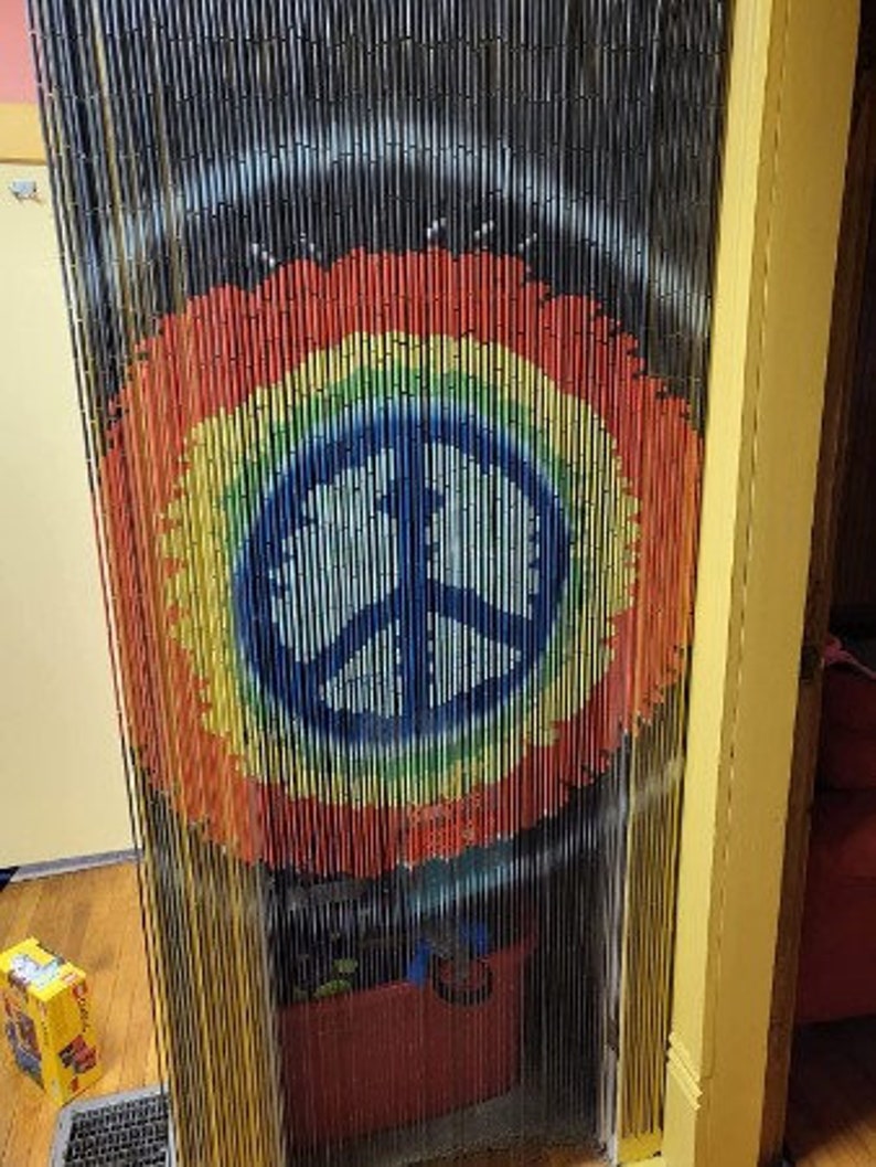 May include: A beaded curtain with a rainbow peace sign design. The curtain is made of black, vertical strands with a large, colorful peace symbol in the center. The peace sign is a circle with a three-pronged symbol inside, painted in blue, yellow, green, orange, and red.