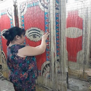 May include: A woman adjusts a beaded curtain made of natural materials. The curtain features a colorful geometric pattern with red, blue, and white accents. The curtain is made of thin, woven strands that create a textured surface.