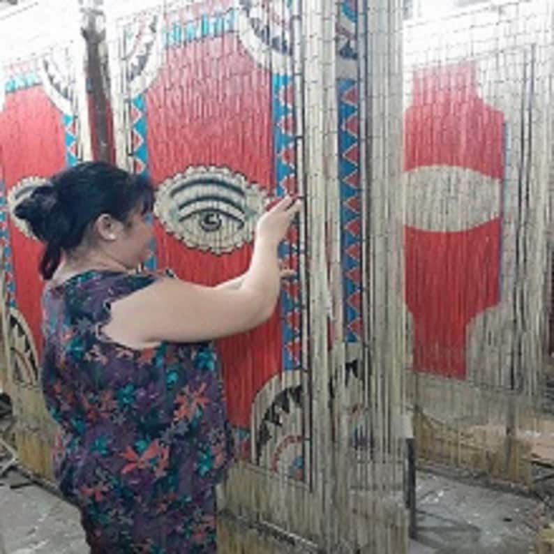 May include: A woman adjusts a beaded curtain made of natural materials. The curtain features a colorful geometric pattern with red, blue, and white accents. The curtain is made of thin, woven strands that create a textured surface.