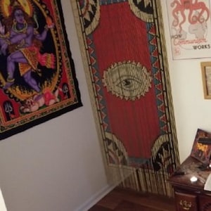 May include: A red and white beaded curtain with a large eye design in the center. The curtain is hanging in a doorway and there are other tapestries and posters on the walls.