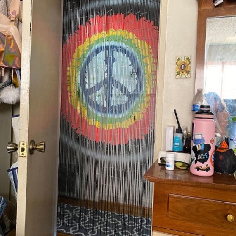 May include: A beaded door curtain with a peace sign design. The peace sign is in the center, surrounded by a rainbow of colors. The curtain hangs in a doorway, with a wooden door frame visible.