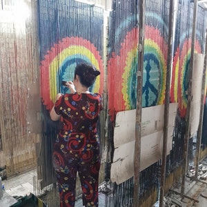 May include: A person is working on a bamboo curtain with a peace sign design. The curtain features a rainbow color scheme with a blue background. The peace sign is in the center of the rainbow design. The person is wearing a red and blue floral print outfit.