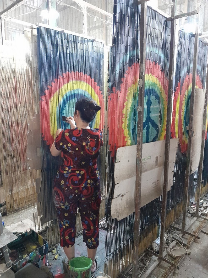 May include: A person is working on a bamboo curtain with a peace sign design. The curtain features a rainbow color scheme with a blue background. The peace sign is in the center of the rainbow design. The person is wearing a red and blue floral print outfit.