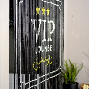 May include: Black beaded curtain with white border and text. The text reads "VIP LOUNGE" with three yellow stars at the top. A fern in a black pot is visible in the lower right corner.